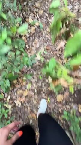 A person in black pants and white shoes walks on a dirt path covered with fallen leaves and some green plants. The camera view is from the person's perspective, looking down at their feet and the path.