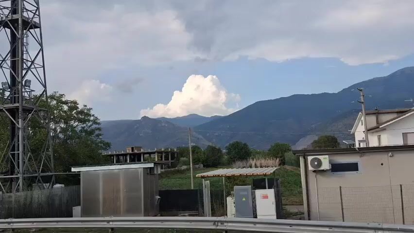 The video shows a cloudy sky above a mountainous landscape. In the foreground, there is a metal tower on the left, a construction site with a building frame in the middle, and a building with an air conditioning unit on the right. There are also some smaller utility boxes and a fence. The overall scene suggests an outdoor, possibly rural or industrial setting.