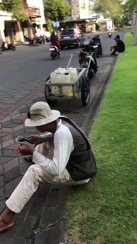 The video pans across a street scene in Boro, East Java. The primary subjects are people and objects, with the setting being an urban street with a grassy median.

In the foreground, a person wearing a white hat and light-colored clothing is sitting on the curb, looking at their phone. They are wearing a dark vest over a long-sleeved shirt and sandals. Next to them, a small, two-wheeled cart with two large plastic containers is attached to a parked scooter.

Further down the sidewalk, another scooter is parked, and behind it, two men in black shirts are sitting on the curb. More scooters are visible parked along the sidewalk, and several are seen riding on the street. The street is paved and has a grassy median with trees and streetlights. Buildings line the opposite side of the street, some with visible signage. One sign appears to read "PHILIPS."

The activity taking place is general street life. People are on their phones, sitting, and riding scooters. The scene suggests a warm, possibly sunny day, given the lighting and the attire of the people. The time of day appears to be late afternoon or early evening, as the sun is casting long shadows. The overall mood is relaxed and casual, depicting everyday life in a bustling East Javanese town. The presence of the cart with containers suggests potential for transport of goods, perhaps water or other liquids, given the containers. The location cues, such as the architecture and the presence of palm trees, are consistent with a Southeast Asian setting, specifically East Java.
