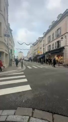 The video shows a street scene in what appears to be a European town, likely in France based on the architectural style. The buildings lining the street are multi-story and have classic French design elements, like shutters and ornate window frames. The sky is overcast. People are walking on the sidewalks and crossing the street at a crosswalk. There are shops along the street, some with displays in their windows. Decorations, possibly for a holiday, are strung across the street. A car is seen driving further down the street. The street is paved, and the edge of the sidewalk is lined with cobblestones. The overall atmosphere is calm and slightly muted due to the weather.