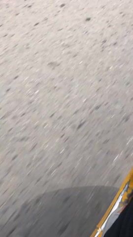 The video shows a low-angle view of a paved road passing under a moving vehicle. The road surface is light gray with scattered dark spots or patches. A portion of the vehicle, likely the frame or side rail, is visible in the lower right corner, painted in yellow with some chipping and wear. The light conditions suggest it is daytime, casting shadows of the vehicle onto the road. The camera is moving forward, following the road as it passes beneath. The road appears to be slightly uneven, creating a subtle shaking effect.