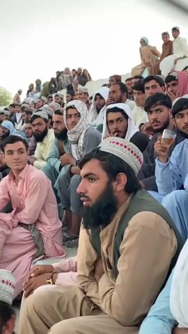 A medium shot captures a large group of men and boys sitting outdoors on tiered seating, likely a stadium or grandstand. The attendees are predominantly dressed in traditional Islamic attire, including various styles of caps and robes in muted colors like beige, light blue, and pink. Many have beards. The lighting suggests it is daytime, possibly late afternoon, with a bright sky. The camera focuses on a few individuals in the foreground, including a man with a beard wearing a light brown vest over a tan shirt and a white and red patterned cap, who is looking to his left. To his left, another young man in a pink robe is looking directly at the camera with a slight smile. The crowd is dense, and most people appear to be looking in the same direction, presumably at an event happening off-camera. The background shows more people seated on higher tiers, and a neutral, overcast sky.