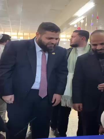 The video captures a scene inside what appears to be a public building, possibly a school or community center. The main subject is a man in a dark suit with a beard, who is the apparent focus of attention. He is surrounded by other men, also dressed in business attire or formal winter clothing. A young girl wearing a hijab and a woman with a lanyard are also present. The man in the suit is gesturing with his hands, as if explaining something. The other men and the girl are looking at him attentively. The background includes decorations hanging from the ceiling, indicating the event may be festive or celebratory. The room has fluorescent lighting and a tiled ceiling. The atmosphere seems friendly and relaxed, with several people smiling and engaging in conversation.