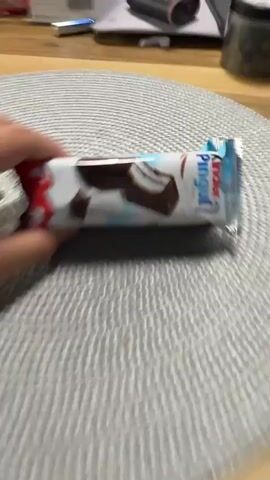 A hand holds a Kinder Pingui chocolate bar in front of a gray circular placemat. The bar is white, brown, and blue. The background is blurry, but there is a white box, a black object, and a jar on a wooden table. The hand moves the bar.