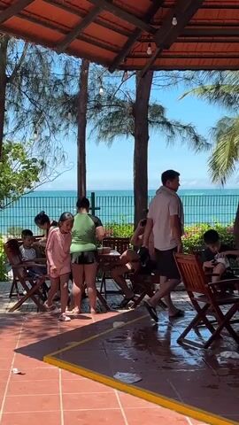 A group of people are gathered under a covered patio, with a beach and ocean visible in the background. In the foreground, a young girl in a pink swimsuit is walking. To her left, a woman in a green shirt and black shorts is standing, and another woman is seated and interacting with a child. To the right of the girl, a man is seated and talking on a cell phone. Further to the right, another child is seated. The patio has red tile flooring and wooden tables and chairs. The sky is blue and clear, and trees are visible.