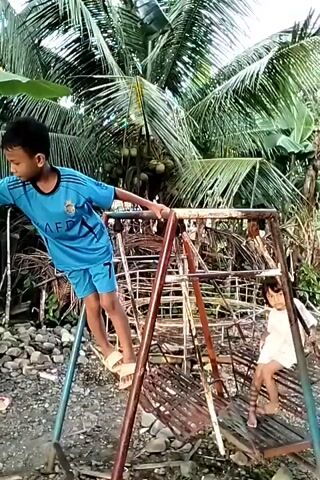 A young boy in a blue shirt and shorts walks across the top of a rusty swing set. A girl sits in the swing. The boy holds on to the top bars of the swing set as he steps from one side to the other. The swing set is in a yard with palm trees and rocks in the background.