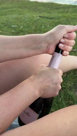 The video shows a close-up of two hands opening a bottle of pink champagne. The bottle has a label that reads "CHEVILLON". The hands are twisting the cork out of the bottle. The scene appears to be outdoors on a grassy area with a body of water in the background. The lighting suggests it is daytime.