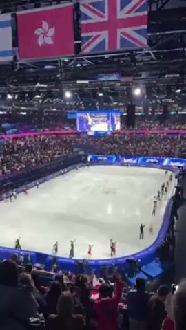 The video shows a large arena filled with people. At center, an ice rink is filled with skaters, most of whom are waving to the crowd. Above the rink, several flags are displayed, including those of the United Kingdom, Hong Kong, and Israel. A large screen shows footage, likely of the skaters. The lighting is predominantly white and blue, and the crowd is cheering. The camera is positioned at an elevated angle, offering a panoramic view of the scene.
