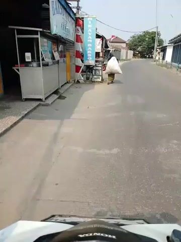 A person is walking down a road, carrying a large white bag over their shoulder. The person is wearing a green dress and a headscarf. The road is paved and lined with buildings on one side. There are several motorcycles parked in front of the buildings. The sky is clear and the sun is shining. The video appears to have been filmed from a moving vehicle, possibly a motorcycle, as the front of a vehicle is visible in the foreground.
