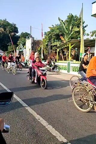 The video shows a street scene with several people riding bicycles and one person on a red scooter. The people on bicycles are mostly wearing casual clothing and some are wearing headscarves. The person on the scooter is wearing a pink shirt and a headscarf. The scooter is carrying two people. In the background, there are trees, buildings, and flags. The sky is clear and the sun appears to be shining. The road is paved and has white lines marking the lanes. The overall impression is of a leisurely group activity or a parade.