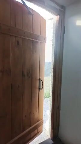 The video opens with a close-up of a wooden door, partially ajar. A black metal handle is visible on the door. As the camera pans slightly to the right, the view through the open doorway reveals a scenic landscape. Rolling hills, covered in dry grass, stretch towards the horizon under a bright, clear sky. In the distance, there are indications of more rugged, mountainous terrain. The ground directly outside the door appears to be paved with interlocking bricks, suggesting an outdoor patio or walkway. A few sparse green plants are visible near the edge of the patio. The lighting indicates it is daytime, likely late morning or early afternoon.