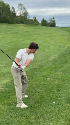 The video shows a man playing golf on a grassy golf course. The man is wearing a white polo shirt, khaki pants, and white sneakers. He is swinging a golf club at a golf ball. The golf course is green with trees in the background. The sky is overcast. The man follows through on his swing, turning to watch the ball’s trajectory.