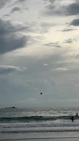 A wide shot captures a beach and ocean under a cloudy sky. In the distance, a small parachute or kite is visible in the air over the water. Waves crash onto the shore, and a rocky outcrop is visible in the ocean. The overall lighting suggests it is either early morning or late evening.