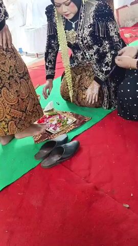 The video shows a wedding ceremony. A woman in a traditional black and gold outfit, adorned with a flower garland, is kneeling. She is being assisted by another person dressed similarly. In front of her is a tray with various items, including flowers and what appears to be food or offerings. Several pairs of black slippers are placed on the floor near her. Another person in a brown and gold patterned skirt stands to her left. The event is taking place on a red carpet with a green mat underneath the tray. In the background, there are white chairs and tables, suggesting an indoor or tented venue. The woman is interacting with someone who is placing their hand on her forehead, possibly a blessing or a ritual. The lighting is bright, indicating daytime.