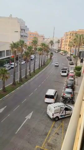 The video shows a street scene with buildings and palm trees lining the road. Several cars are parked along the side of the road and are driving on the road. There are also a few motorcycles. The sky is overcast.