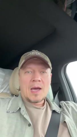 A man is sitting in the driver's seat of a car, singing. He is wearing a khaki baseball cap, a light green jacket, and a beige t-shirt. He has light skin, blue eyes, and short blond hair. The interior of the car is modern with a dark gray ceiling and tan leather seats. A white sheet is draped over the passenger seat. The man is looking directly at the camera as he sings, then looks slightly to his left and then back to the camera. He is wearing a seatbelt.