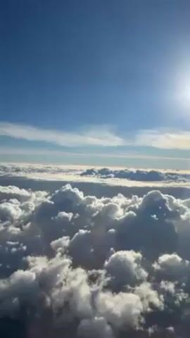 The video shows a view from an airplane window above a sea of puffy white clouds under a clear blue sky with a bright sun. There is no person visible in the video. The airplane is flying, as indicated by the clouds moving. A voice is heard speaking in Italian.