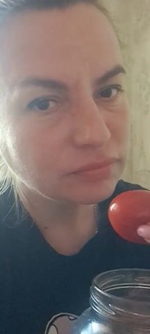 A fair-skinned woman with light brown hair pulled back in a low ponytail is shown in a close-up video, where she picks a red tomato from a glass jar and attempts to put it in her mouth. She’s wearing a black t-shirt and has long, light pink painted nails. Her face is close to the camera, highlighting her features, including her slightly pursed lips. The tomato is positioned between her lips, and she uses her index finger to push it further. The background is out of focus, but appears to be an indoor setting.