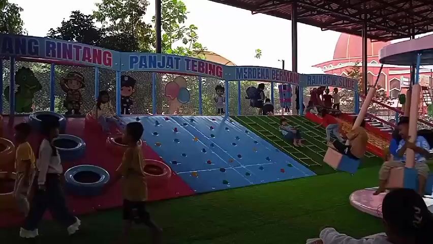 The video shows a group of children playing in a playground with various activities. The playground has a colorful structure with climbing walls, slides, and a rope ladder. The structure is labeled with the words "OBANG RINTANG," "PANJAT TEBING," "MERDEKA BERMAIN," and "RAINBOW SLIDE." Some children are climbing, sliding, or walking around on the artificial grass. In the background, there is a building with a red dome. The playground is covered by a metal roof.