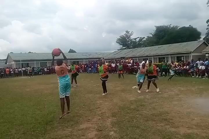 The video shows a netball game being played outdoors. Several female players wearing different colored uniforms are actively engaged in the game on a grassy field. One player in a light blue uniform is holding the ball, while others are moving around, possibly trying to intercept or position themselves for a pass. In the background, a crowd of people, including many students, are seated and watching the game. Buildings with multiple windows line the background, along with some trees and an overcast sky.