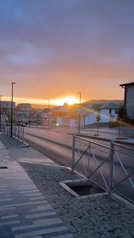 The video shows a street scene at sunset. The sun is visible on the horizon, casting a warm glow over the sky and landscape. Buildings line the street, and there are streetlights and a sidewalk. The road is mostly empty, with a few cars visible in the distance. The overall impression is one of a peaceful and quiet evening.