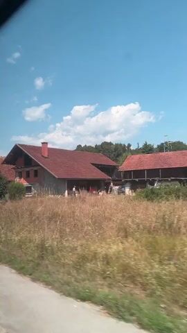 The video appears to be filmed from a moving vehicle, looking out a window. The scene depicts a rural or suburban area with several houses visible. The houses have red tile roofs and appear to be of varying sizes and styles. There are trees and vegetation surrounding the houses, including what looks like dry grass in the foreground. The sky is blue with some white clouds. In some frames, there are glimpses of people and cars. The overall impression is of a sunny day in a residential area.
