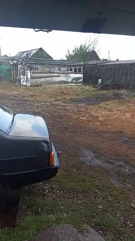 This video shows a black car parked on a muddy surface with some green grass. In the background, there are several wooden structures, some of which appear dilapidated or under construction. There is also a tractor visible in the distance. The sky is overcast, and it appears to be raining or has recently rained, as evidenced by the wet surfaces and water dripping from the roof of the structure the camera is under. The overall scene depicts an outdoor area, possibly a rural yard or a farm, with a focus on the car and its surroundings.