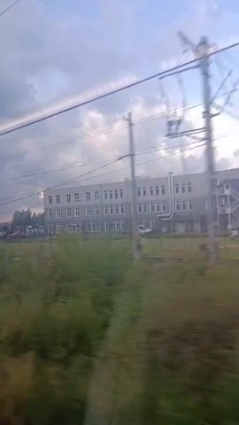 The video shows a view from a moving train, looking out the window at a cloudy sky and a landscape. The landscape includes a grey multi-story building with many windows, a field of green grass and weeds, a set of train tracks, and what appear to be power lines running across the sky. In the background, there are several vehicles including cars, trucks, and possibly a construction site with a large blue shipping container. The train passes by a red locomotive on the tracks and then another train with silver carriages. The view is slightly blurred due to the movement of the train.