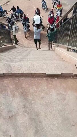A view looking down a flight of concrete stairs, beside a metal railing, shows multiple men on motorcycles parked at the base of the stairs, some holding white buckets. A young person wearing shorts and carrying a white bucket on their head walks down the stairs toward the motorcycles. Another young person walks down the stairs at the same time. The ground around the stairs and motorcycles appears to be dirt. The scene is brightly lit.