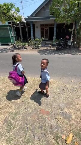 Two young children, a girl and a boy, are standing on a dirt path next to a paved road. The girl, wearing a school uniform with a blue and white collared shirt, navy skirt, and white socks with sneakers, carries a large pink backpack. The boy, also in a school uniform consisting of a blue and white shirt and navy shorts with sneakers, faces the girl and extends his hand. The girl turns away from the boy and runs across the paved road, her backpack bouncing. The background shows a house with a dark tiled roof, a motorcycle parked in front, and trees. The scene is outdoors on a sunny day, with a dry, grassy area on the side of the road.