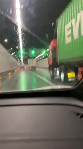 The video shows a view from inside a car driving through a tunnel at night. The windshield of the car has water droplets on it, distorting the view slightly. The tunnel is lit by overhead lights, some of which are white and some are green. There are other vehicles in the tunnel, including a red truck with a green container on its trailer in the right lane and a white truck with a container in the left lane. The road surface appears wet. The video captures the movement of the car through the tunnel, passing the other vehicles.