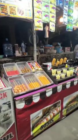The video pans across a food stall at night. The stall is brightly lit with overhead lamps and displays a variety of food items. In the foreground, there are shelves filled with packaged snacks and condiments. Towards the center of the stall, there are several small containers holding what appears to be chopped food, and a display of corn dogs. Further back, there are more packaged goods, bottles of drinks, and a grill with food cooking on it. Two women are working at the stall, one preparing food and another attending to customers. The background shows a dark, outdoor environment, likely a street or market. The stall is decorated with colorful banners and images of food.