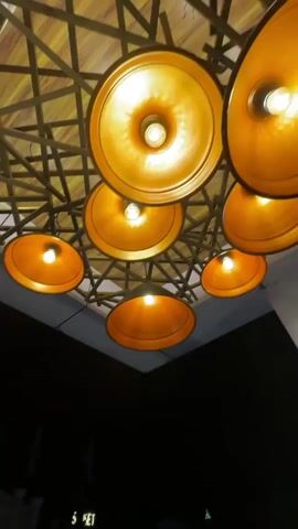 The video pans down from a group of warm-toned, circular pendant lights suspended from a latticed ceiling to reveal a nighttime urban scene. Below the roof of the building where the camera is located, buildings with multiple floors and various architectural styles line the street. Light emits from windows and illuminated signs. The sky is dark.