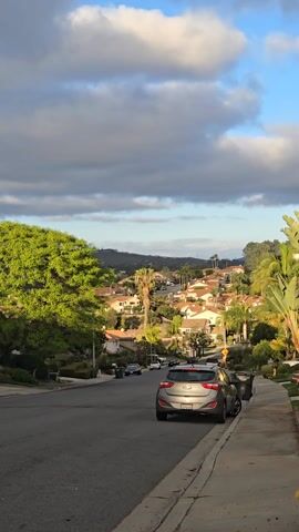 The camera shows a residential street scene. A gray hatchback car is parked on the right side of the road, facing downhill. The street slopes downwards, providing a view of houses with red tile roofs nestled among green trees, including palm trees, on the hillside. A distant mountain range is visible under a sky with scattered clouds. The lighting suggests it is daytime, possibly late afternoon. There is no visible human activity in the scene.