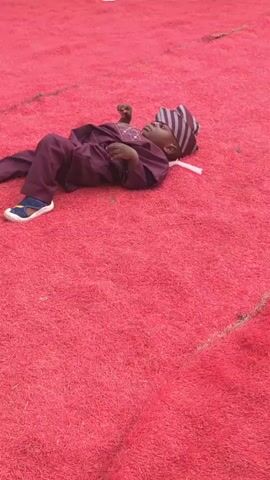 A young boy wearing a maroon outfit with a striped hat and sandals lies on a red carpet. He moves slightly and looks around. The carpet is fluffy and covers the ground. In the background, white tablecloths and chairs are partially visible. A rhythmic beat plays in the background, and someone can be heard saying something in a low voice.