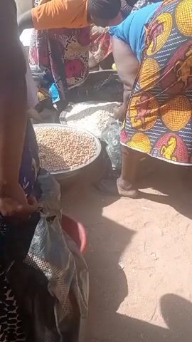 The video shows three women in an outdoor marketplace handling food. The woman on the left, wearing an orange shirt, uses a green scoop to transfer tan, irregularly shaped pieces of food from one metal pan to another. In front of her is a large metal pan filled with whole, round, tan beans or nuts. The woman on the right wears a colorful, patterned dress with geometric and horse-like shapes. A third woman wearing blue stands behind them. All three are standing on what appears to be packed sand. There is a black plastic bag partially visible.