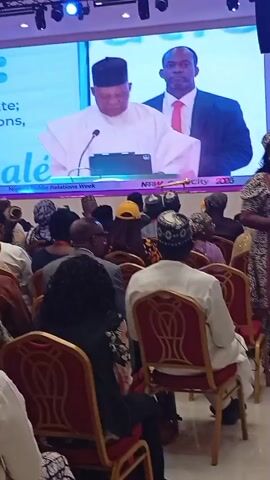 The video depicts an indoor public speaking event or conference held in Tudun Nupawa, Nigeria. A large screen at the front of the venue displays two men. One man, dressed in a white agbada and a black cap, is speaking at a podium with a microphone and a tablet device. The second man, wearing a dark suit and a pink tie, stands behind him.

The screen also shows partially visible text like "te;", "tions,", and "alè," along with "Nigeria Public Relations Week" and "NRRW City 2026," indicating the nature and likely future host city of the event.

In the foreground and middle ground, a diverse audience is seated in rows of red chairs, facing the screen. Most attendees are seen from behind, and many are wearing traditional Nigerian attire or head coverings. One woman in a patterned black and white dress walks across the frame from right to left, between the camera and the seated audience. The atmosphere appears attentive, with the audience focused on the presentation displayed on the large screen.