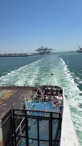 The video shows the view from the back of a moving ship on a sunny day. The ship is leaving a white wake behind it in the blue water. In the distance, there is land with several large cranes. The deck of the ship is visible, with a metal grate and some equipment. The sky is clear and blue.