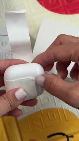 A person with manicured fingernails is opening a new white case for AirPods. The case is held against a background of white boxes and a colorful, childlike mat. Inside the case are the AirPods themselves, also white. The case’s light is on and glowing orange.