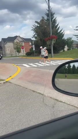 Two people, a man and a woman, wearing white hats, are walking across a crosswalk. The woman is wearing a white tank top and black shorts, and the man is wearing a grey t-shirt and grey shorts. They are walking from left to right in the frame. A black minivan is stopped at the crosswalk, waiting for them to pass. The scene takes place on a cloudy day, with houses and trees in the background. The perspective is from inside a vehicle, looking out through the passenger-side mirror, which partially obscures the view of a single person walking in the distance.