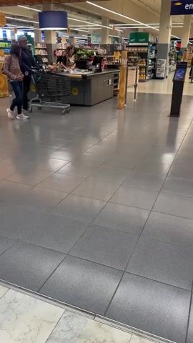 A middle-aged couple walk through a brightly lit supermarket. The man pushes a silver shopping cart, and the woman walks beside him. The woman wears a floral shirt, jeans, and pink sneakers. She has a small green backpack. The man wears a striped shirt and dark pants. The floor is gray tile. Aisles with various products are visible in the background. A black kiosk stands to the right of the couple. The couple walks from left to right in the frame. The store's exit sign is visible in the upper right corner of the frame.