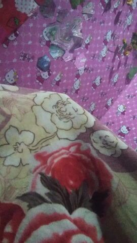 A young woman is lying on a bed indoors in Kecamatan Cimaung, Indonesia. She is wearing a red top and is partially covered by a floral patterned blanket featuring red, cream, and brown flowers. Her head rests on a pillow with a pink Hello Kitty pattern, and the bed is also covered with a pink Hello Kitty patterned sheet. Scattered on the Hello Kitty sheet are several small, clear, faceted objects. The woman looks towards the camera with a slight, calm smile. The camera briefly shows a close-up of the floral blanket and then blurs before settling on the woman's face.