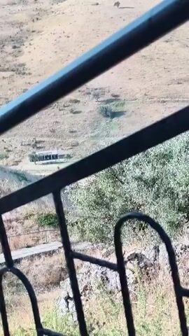A video captures an outdoor landscape. The foreground features a black metal railing, suggesting a balcony or staircase. Beyond the railing, a dry, grassy hillside unfolds, dotted with sparse trees. In the distance, rolling hills stretch towards a hazy horizon under a clear sky. The scene appears to be a rural or natural environment.