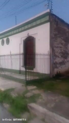 The video shows a street in a town or city at dusk or dawn. The street is paved and runs alongside a long, single-story building painted white with a green trim. The building has several arched doorways, some of which appear to be entrances. There are also two circular windows along the top of the building. A black, wrought-iron fence runs along the front of the building, separating it from the sidewalk.

The street is mostly empty, except for a few parked cars and a couple of people in the distance. There are streetlights illuminating the road. Overhead, power lines crisscross the sky. A tree is visible on the left side of the road.

The camera appears to be moving, giving the impression of traveling down the street. The video is slightly shaky.