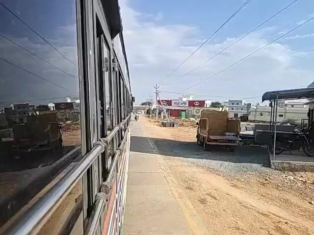 This is a first-person perspective video taken from inside a moving vehicle, likely a bus, with a view of the right side of the vehicle and the road ahead. The vehicle appears to be traveling on a paved road through a semi-urban or developing area. The sun is shining, indicating a bright, daytime scene with a clear blue sky and scattered clouds.

On the right side of the road, there are piles of dark, rectangular stones, possibly marble or granite slabs, suggesting a stone quarry or a construction material storage area. Further down the road, there are buildings under construction or recently built, appearing to be a mix of residential and commercial structures. Some of these buildings have red and white signage. There are also utility poles with overhead wires running along the road.

A person is seen riding a motorcycle on the road, traveling in the same direction as the camera vehicle. A small, light-colored truck with a flatbed carrying some wooden crates or materials is parked on the right side of the road. The ground on the sides of the road is dusty and appears to be unpaved in some areas. The reflection on the window of the vehicle shows the surroundings, including the sky and some buildings.