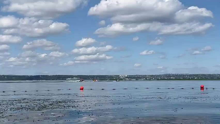 A wide shot shows a calm body of water with a distant shoreline under a partly cloudy blue sky. In the foreground, a line of red buoys are visible just below the surface of the water, with a few pieces of debris floating on the surface. Several jet skis are being ridden in the middle ground, creating wakes. The distant shoreline features trees, buildings, and a bridge. The overall scene suggests a recreational activity on a lake or bay.
