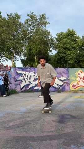 A man is skateboarding in an outdoor skate park. He is wearing a short-sleeved, collared button-down shirt, black pants, and black shoes. He performs an ollie, and briefly loses his balance before regaining control. The ground has painted designs. Behind him is a wall with graffiti. Other people are in the background, one riding a scooter. Trees are also visible in the background. The sky is visible and light in color.