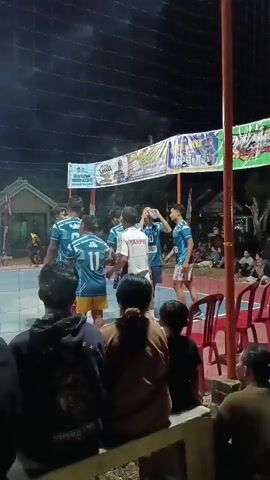 The video shows a group of young men playing volleyball at night. They are wearing athletic uniforms. There is a crowd of spectators watching the game from the sidelines. The court is lit by overhead lights, and there are banners and signs in the background. The atmosphere appears to be lively and engaged.