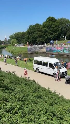 A bright, sunny day scene unfolds by a river. In the foreground, a lush green bush borders a paved pathway. People are seen walking along the pathway and on the grass beside the river. A white van is parked on the grass near the pathway, with a family, including a child in a stroller, near it. Further down the riverbank, several tents and stalls are set up. Some people are gathered near these tents, while others are in or near the water. Kayaks and canoes are visible on the river. A dark-colored station wagon is parked on the grass near one of the stalls, identified by a sign as "RIVER CASINO". More people are scattered along the opposite bank of the river, which is lined with trees. The overall atmosphere appears to be a public gathering or event near a river.