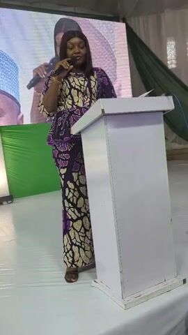 A woman stands at a podium, speaking into a microphone, wearing a purple and white patterned dress. The room is decorated with white and green drapes, and artificial grass covers the floor. There are people in the background, some seated, others standing, and a videographer with a camera on a tripod. A large screen is visible behind the speaker, displaying an image.